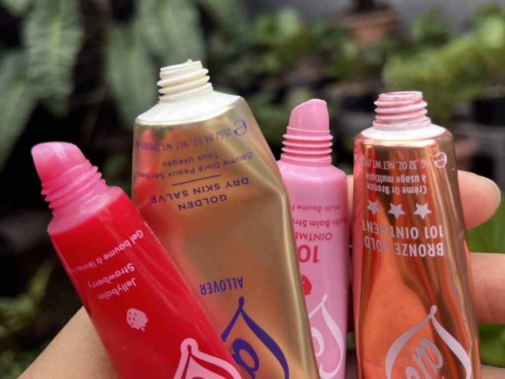 Close-up of Lanolips balm applicators from different product lines held by hand against a blurred green garden background.