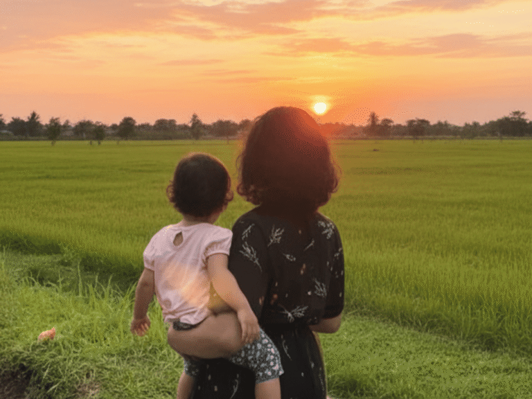 A mother carrying her daughter while watching the sunset over a green field.